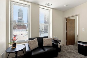 Una sala de estar con un sofá de cuero negro, dos mesitas auxiliares, una pequeña planta en maceta y vistas a un paisaje urbano a través de amplios ventanales.
