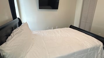 Un dormitorio con cama, televisor de pantalla plana montado en la pared, armario con puertas persianas y ventana con cortinas.