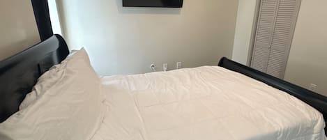 A bedroom with a bed, a flat-screen TV mounted on the wall, a closet with louvered doors, and a window with curtains.