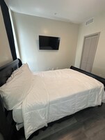 Un dormitorio con cama, televisor de pantalla plana montado en la pared, armario con puertas persianas y ventana con cortinas.