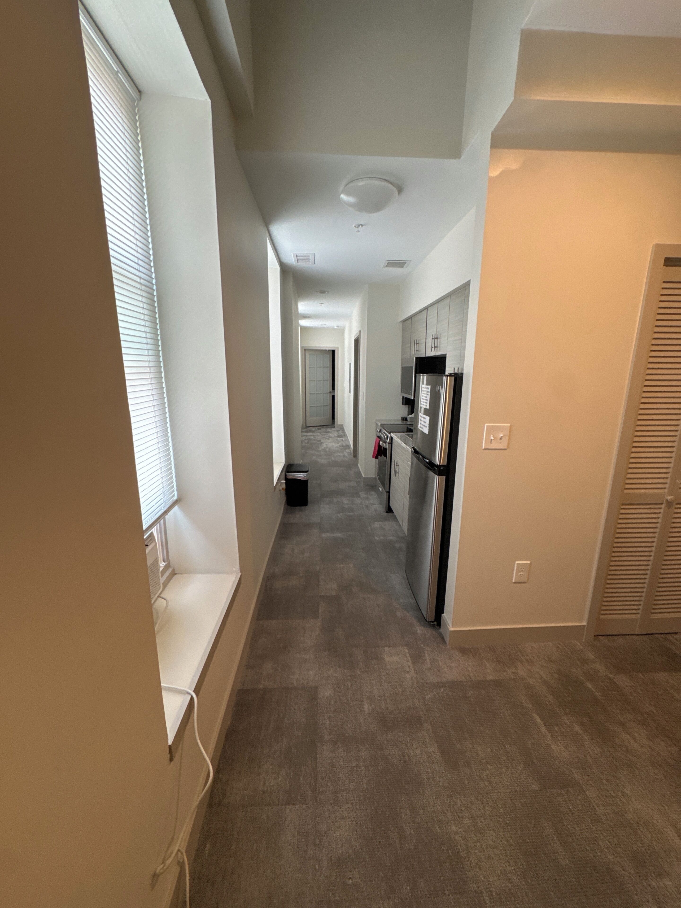 A narrow hallway with a kitchen area on the right, a trash bin, and a door at the end.