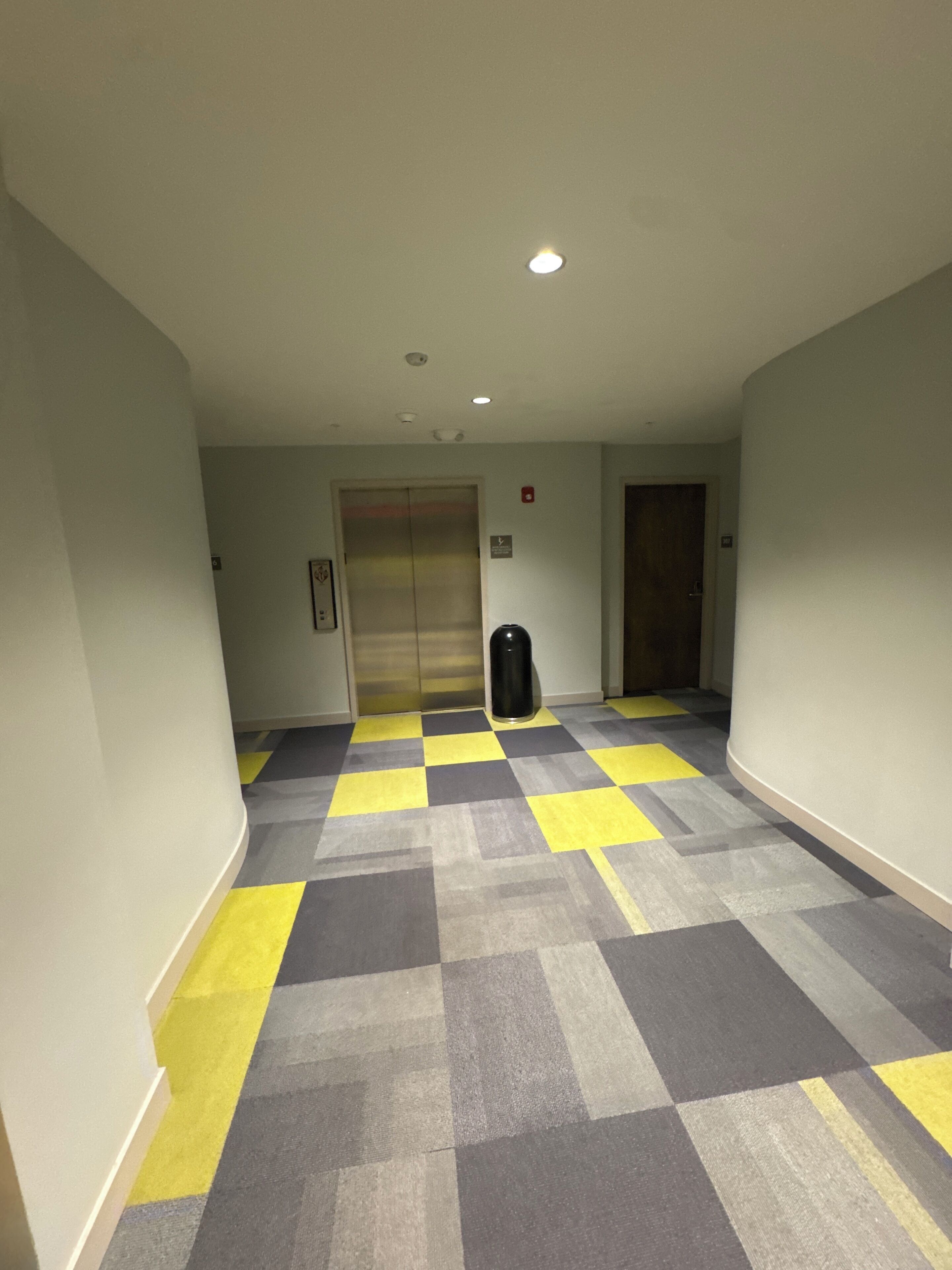 Um corredor com padrão de piso xadrez, um elevador e uma lixeira.