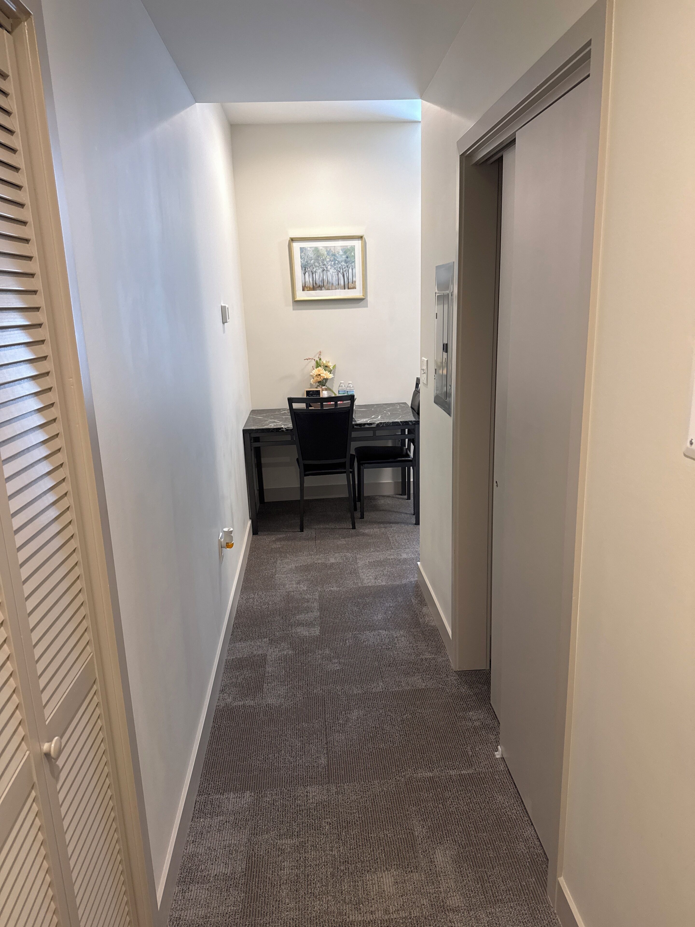 A narrow hallway with a carpeted floor, a door on the right, and a desk with a chair and a vase of flowers in the background.