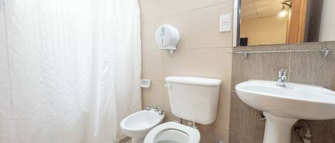 A bathroom with a shower curtain, two toilets, a bidet, a sink, and a hand dryer.