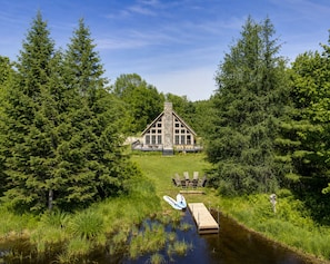 Un chalet doté d’une cheminée en pierre, entouré d’une forêt dense, avec un quai et un bateau, ainsi que des sièges à l’extérieur.