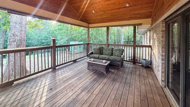 A covered porch with a wooden deck, a cushioned seating area, and a view of the surrounding forest. - Peaceful country home minutes from Lake Mitchell (Clanton)