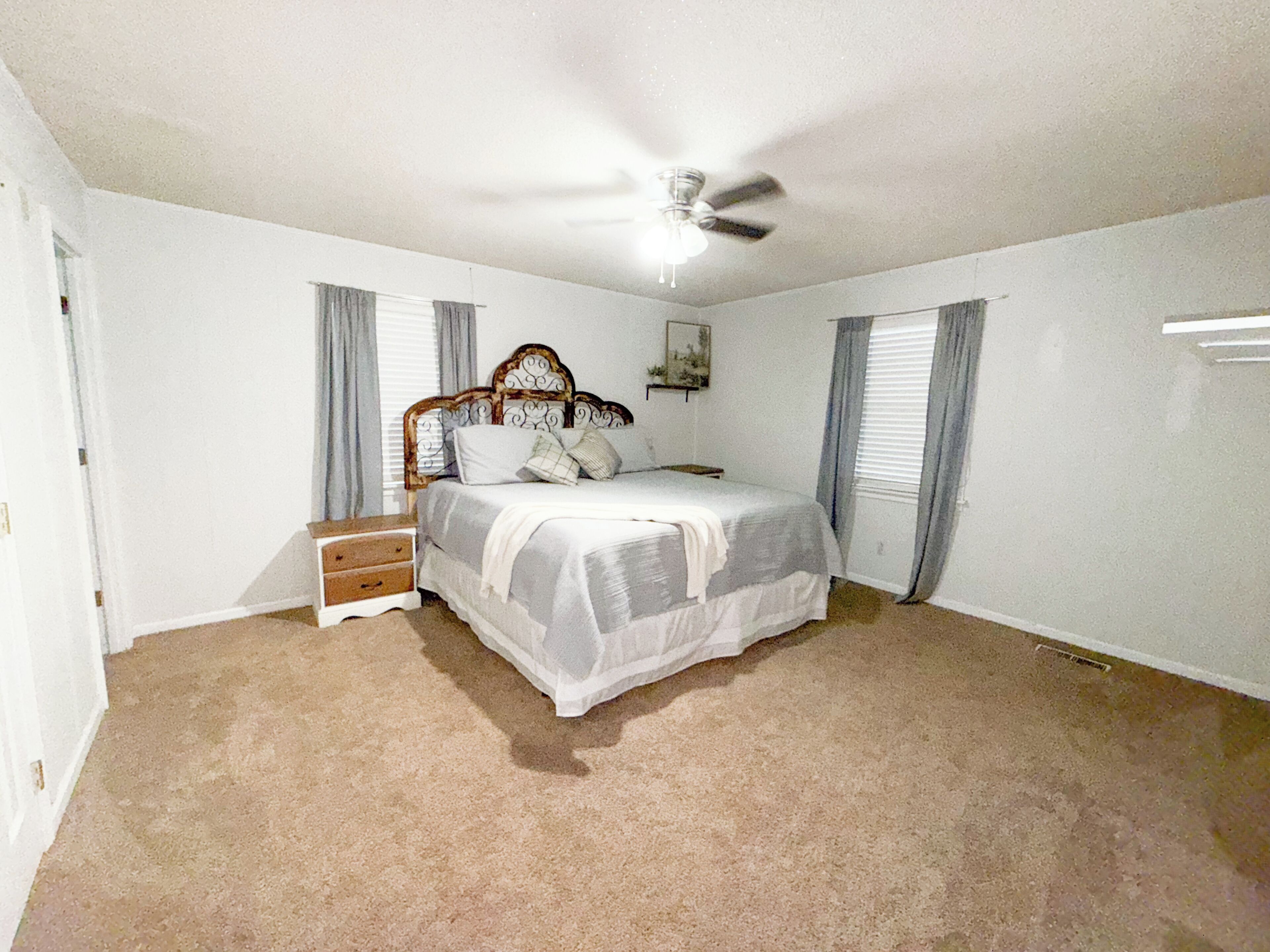 Une chambre à coucher avec un lit, une table de chevet, un ventilateur de plafond et une fenêtre avec des rideaux.
