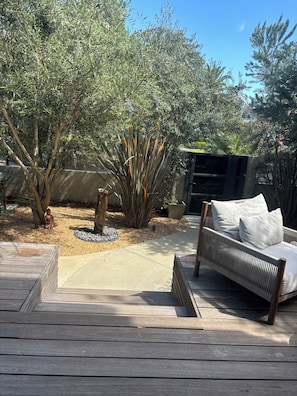 Eine Terrasse mit Holzdeck, einem weißen, gepolsterten Outdoor-Sofa und verschiedenen Pflanzen, darunter ein Olivenbaum und eine hohe, schlanke Pflanze mit langen Blättern.