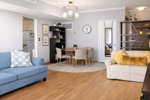 A living room with a blue sofa, a white sofa, a dining table with chairs, a clock on the wall, and a bookshelf. - Oceanfront Paradise Suite - Ocean, Pool & Gym (Hollywood)