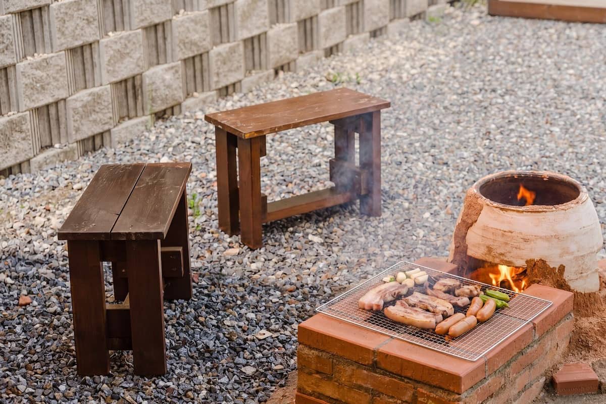 A barbecue grill with sausages and vegetables, a wooden table, and a fire pit.