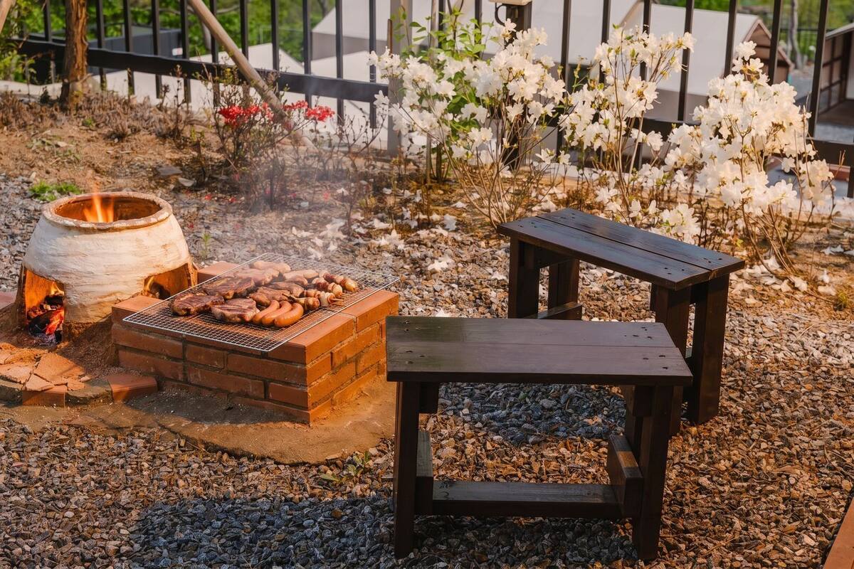 A barbecue grill with food, a fire pit, and a wooden bench.