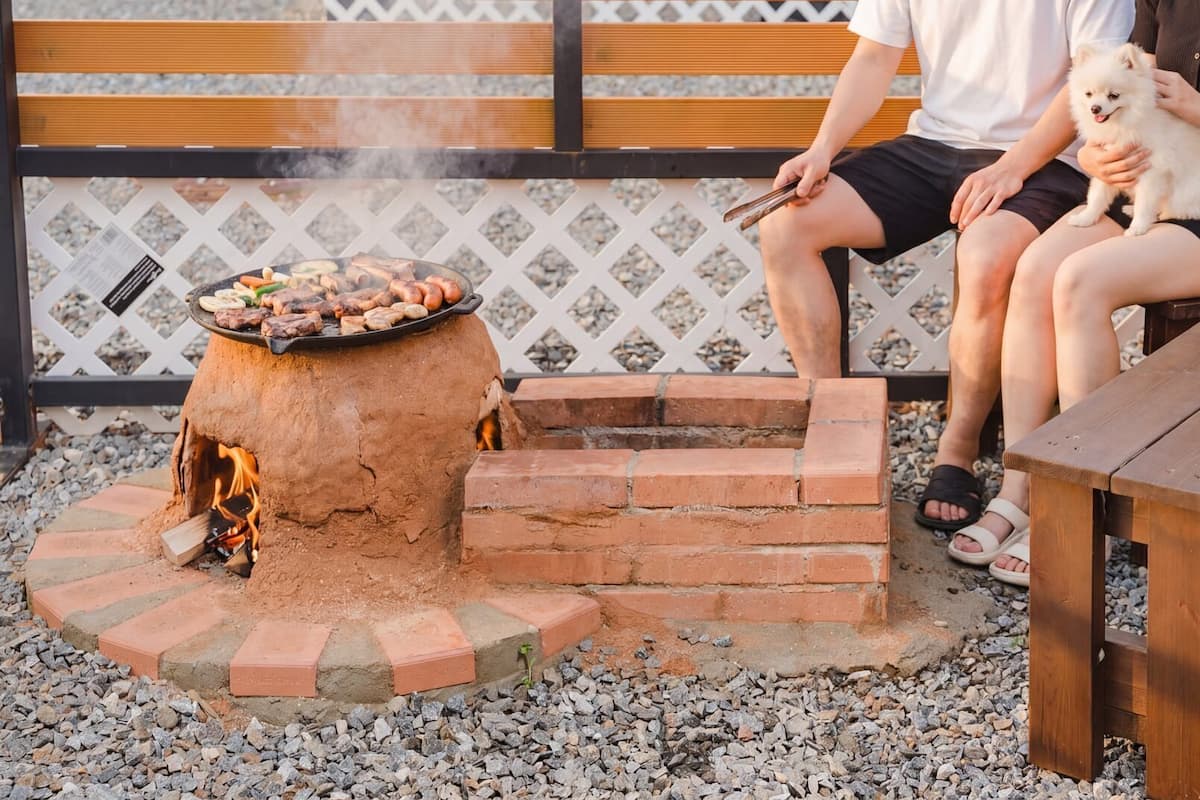 A person grilling outdoors with a small fire pit and a wooden table nearby.