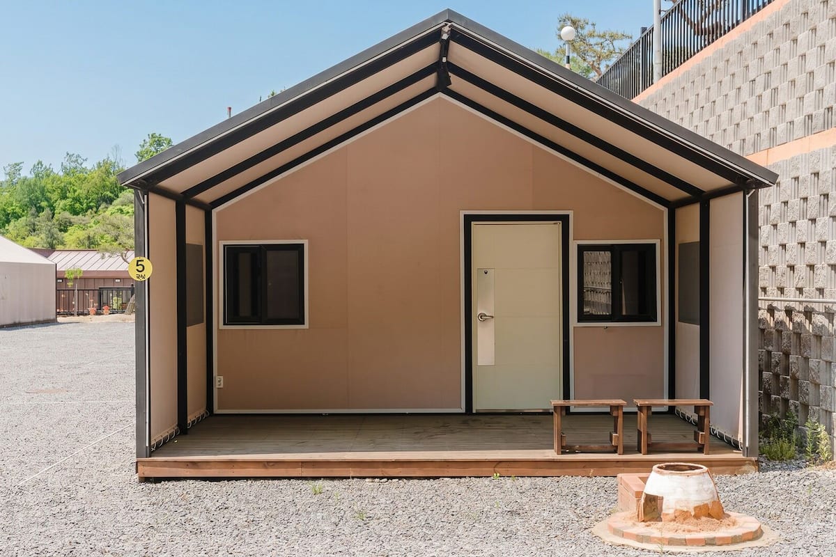 A small, single-room structure with a door, two windows, and a bench outside.