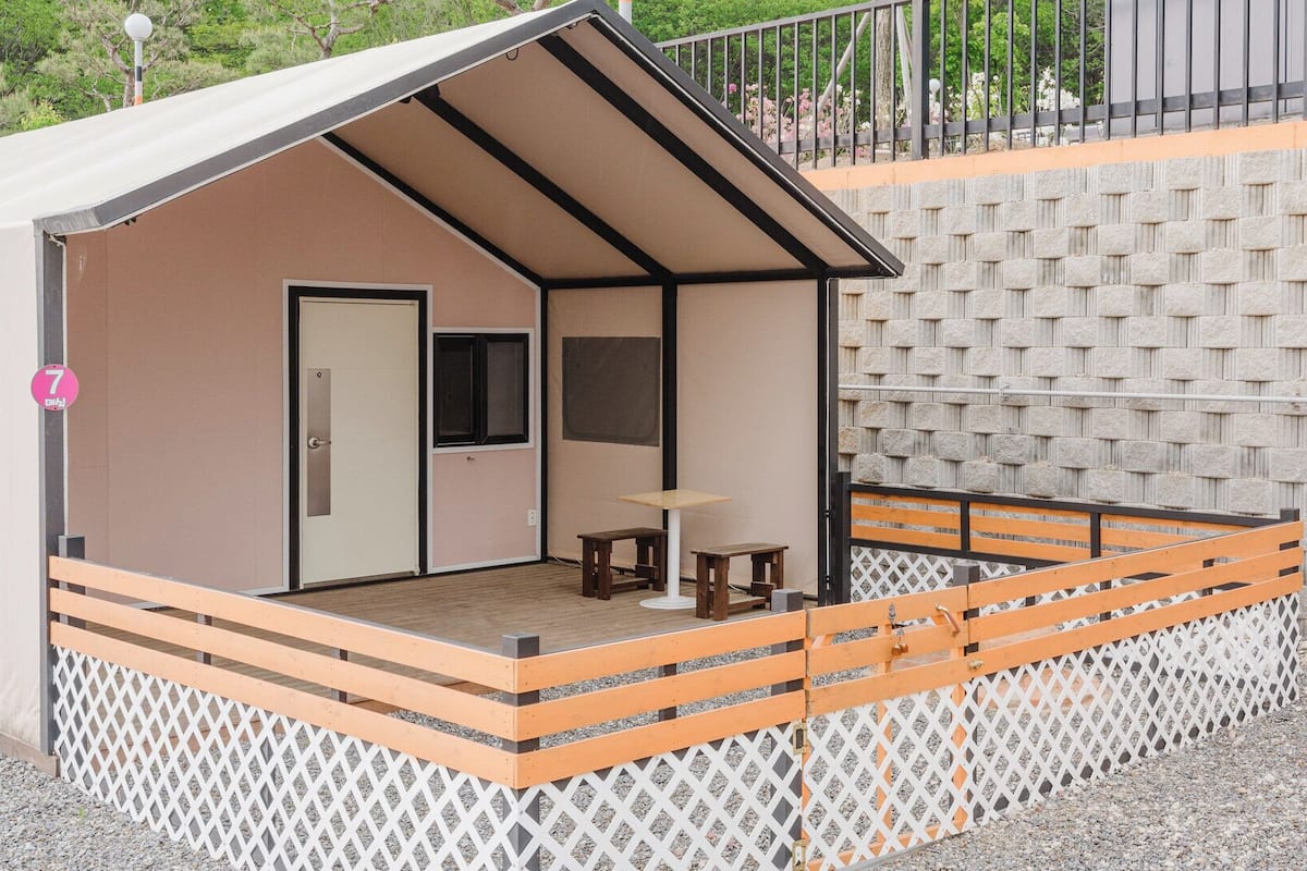 A small, single-room cabin with a sloped roof, a door, a window, and a small table and chair set outside.