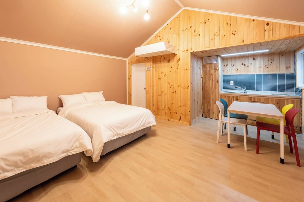 A bedroom with two beds, a wooden wall, a window, and a small kitchen area with a table and chairs.