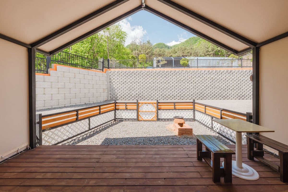 A room with a sloped ceiling, a wooden bench, a table, and a view of a garden with a brick wall and a fence.