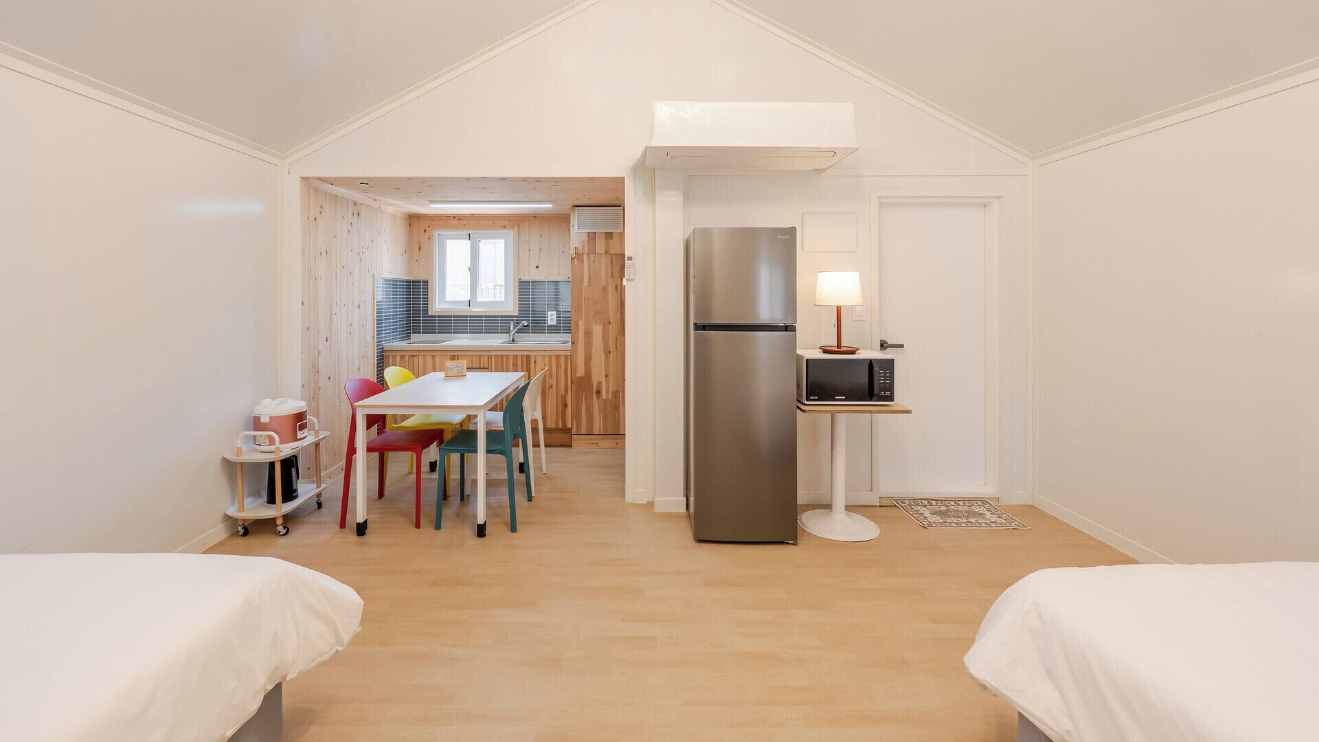 A compact room with two beds, a kitchenette, a dining table with colorful chairs, and a refrigerator.