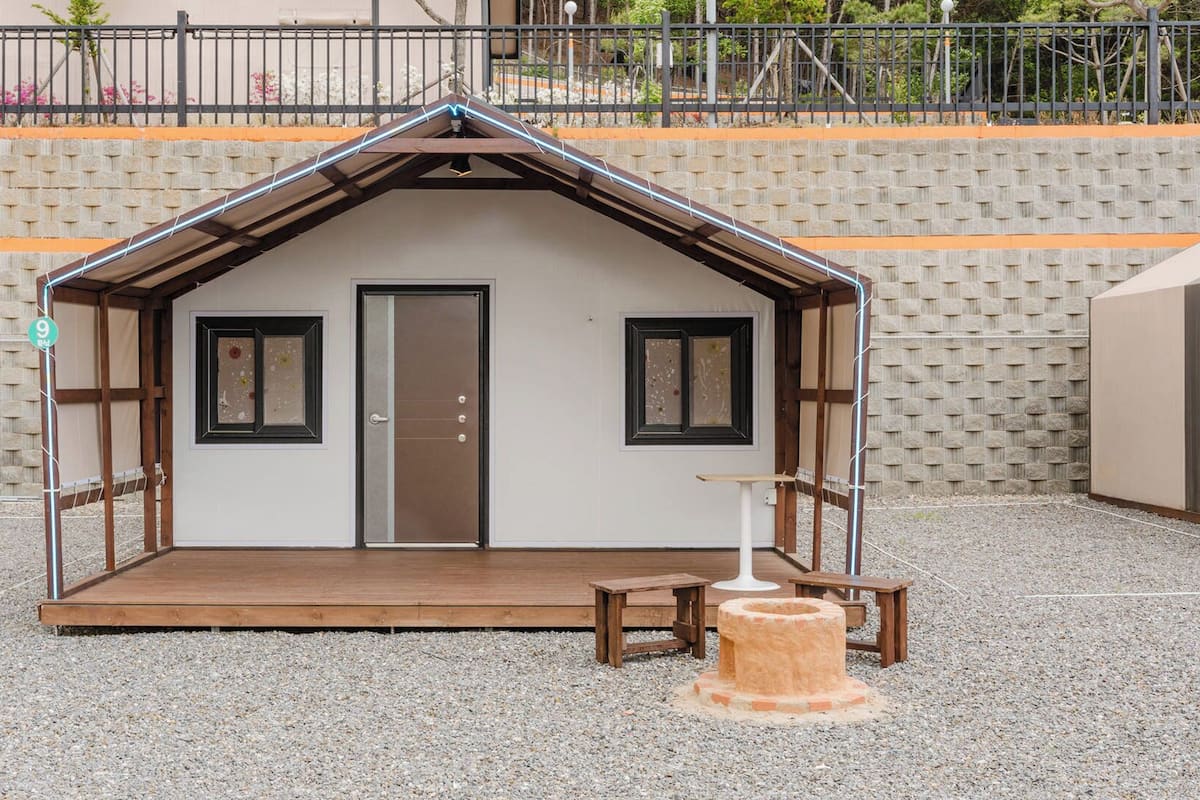 A small, single-room structure with a sloped roof, a door, two windows, a bench, and a small table.