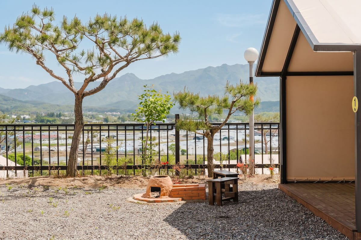 A balcony with a view of mountains, a small tree, a brick oven, and a table.
