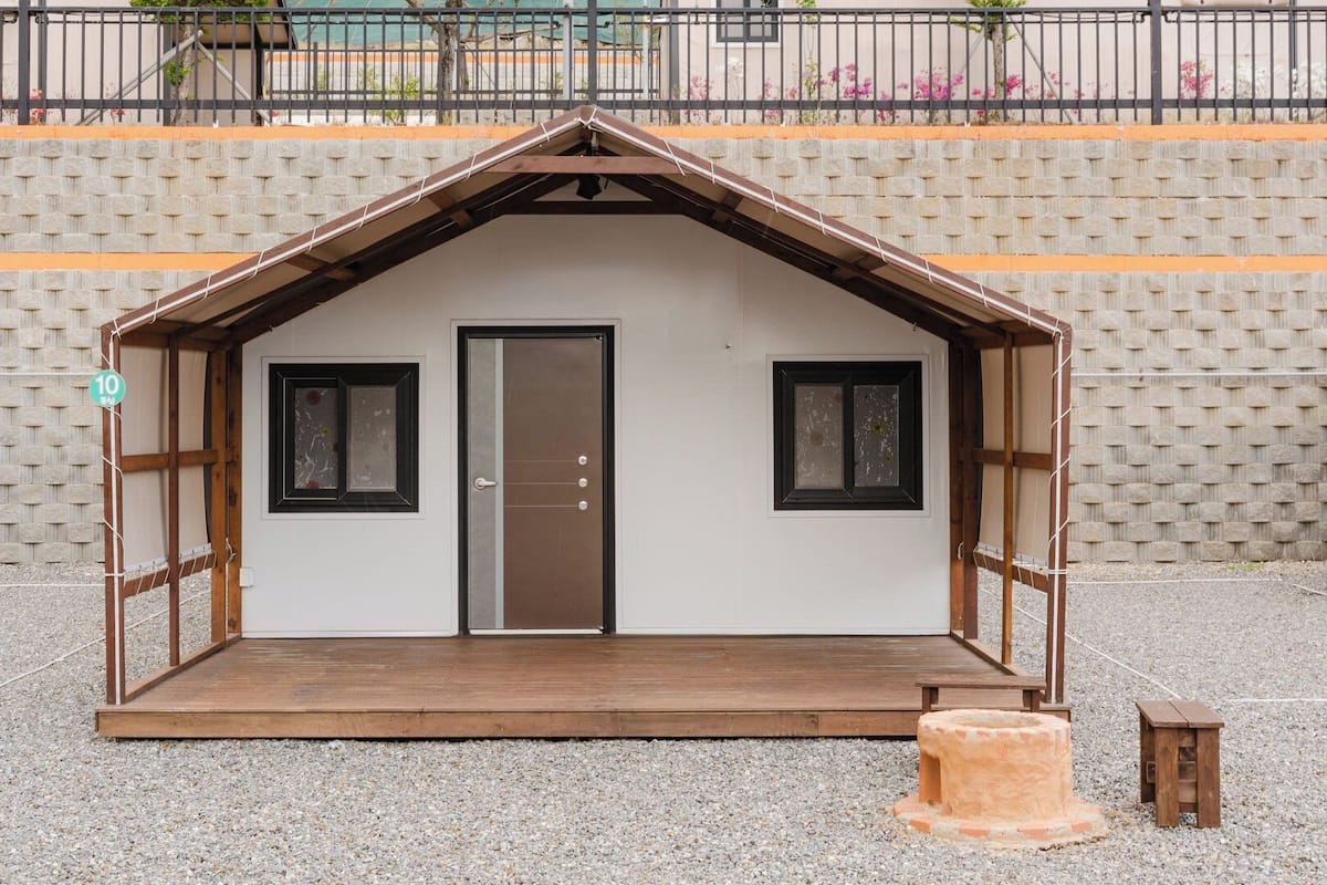 A small, single-room structure with a door and two windows, a wooden deck, and a small stool.