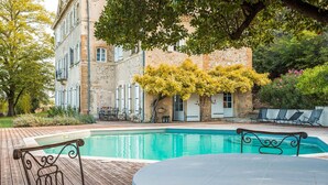 A poolside area with a table and chairs, a stone building with a door, and a tree with yellow leaves. - Château avec piscine privée (Valréas)