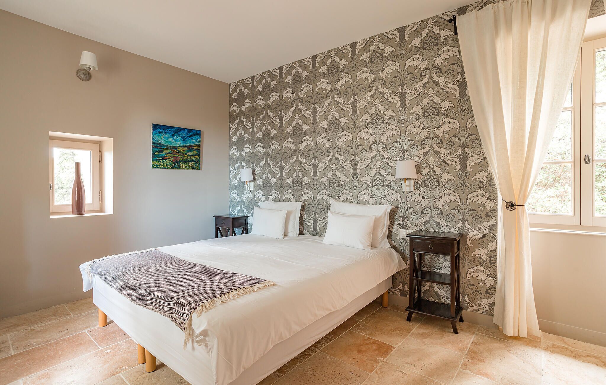 Une chambre à coucher avec un grand lit, deux tables de chevet, une fenêtre avec des rideaux, une lampe fixée au mur et un tableau.