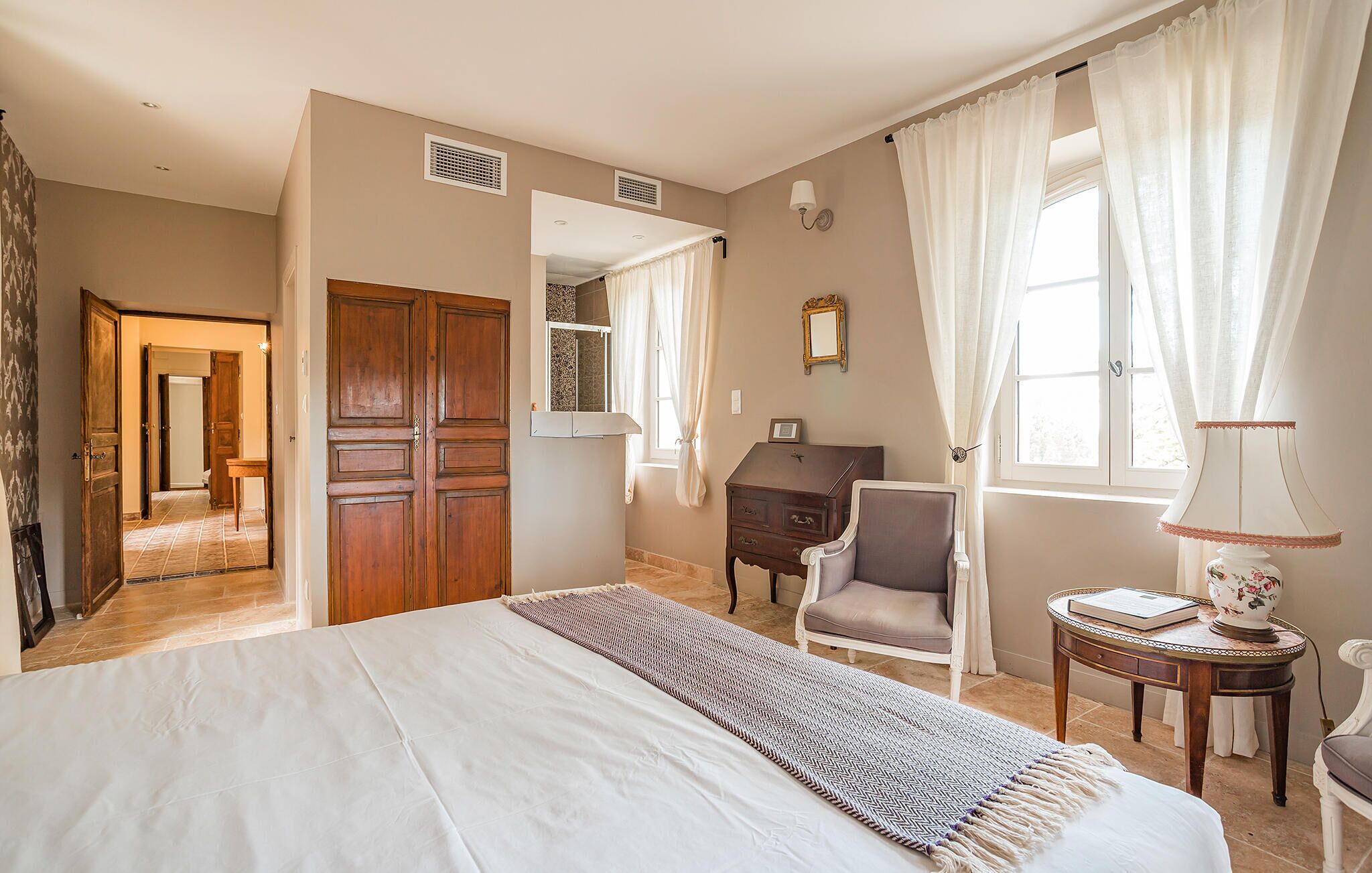 Ein Schlafzimmer mit Bett, Stuhl, Kommode, Lampe und einem Fenster mit Vorhängen.