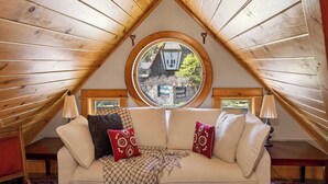 Una stanza accogliente con soffitto spiovente, una finestra tonda, un divano con cuscini decorati e due lampade.
