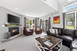 A modern living room with a fireplace, leather furniture, a flat-screen TV, and large windows. - Wildflower Townhouse, Steps to Holiday Valley (Ellicottville)