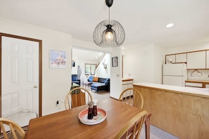 A dining area with a wooden table, chairs, a decorative plate with salt and pepper shakers, a modern pendant light, and a kitchen area with cabinets and a refrigerator.