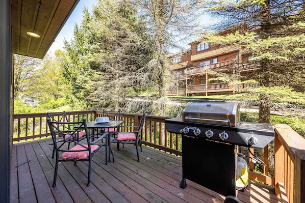 A deck with a grill, chairs, and a view of a cabin.