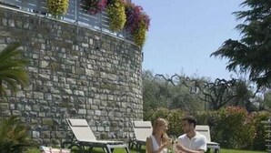 Una piscina con dos personas sentadas al borde, un muro de piedra y mobiliario de exterior.