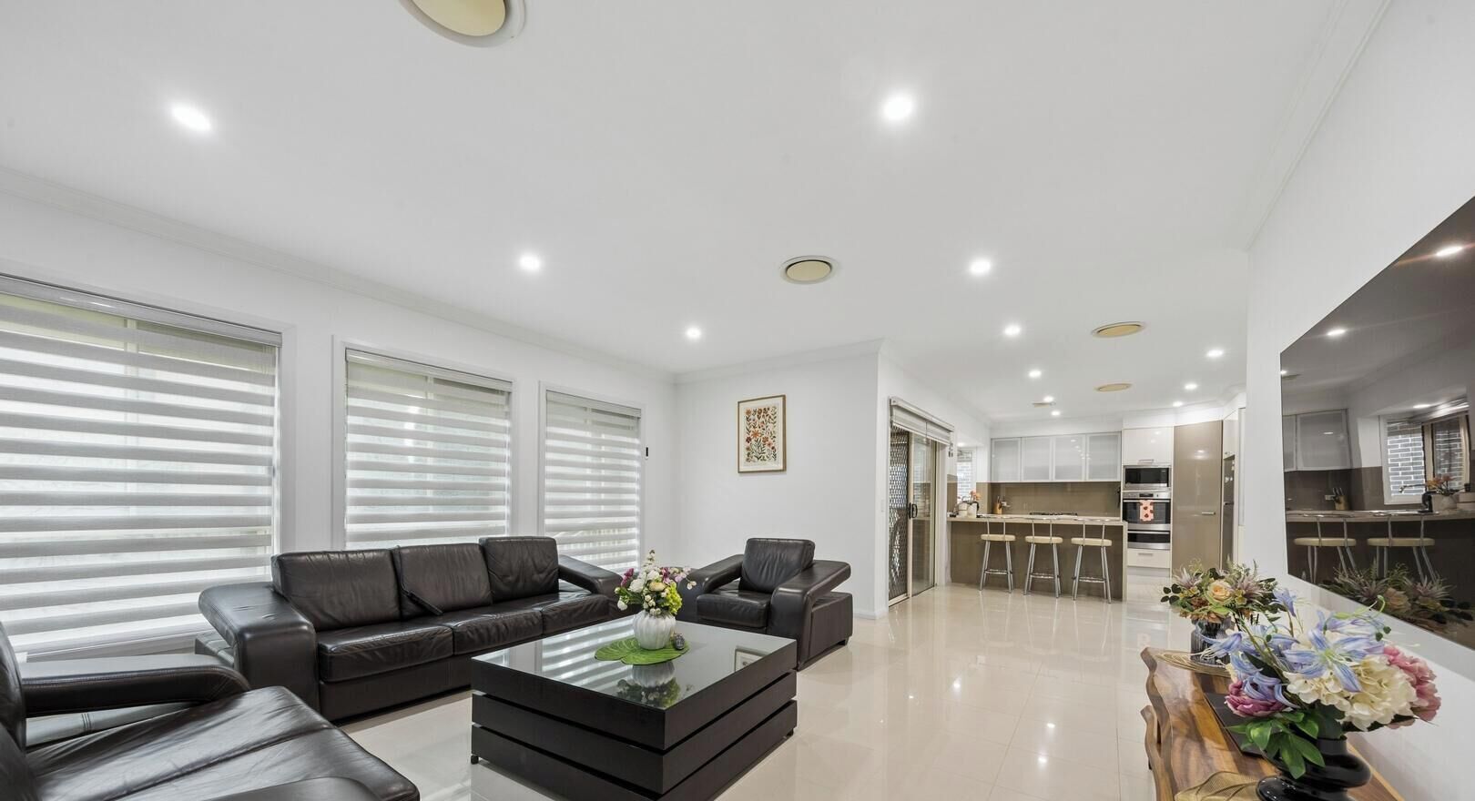 A modern living room with a black leather sofa, a glass coffee table, and a kitchen area in the background.