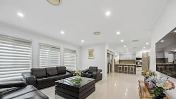 A modern living room with a black leather sofa, a glass coffee table, and a kitchen area in the background.