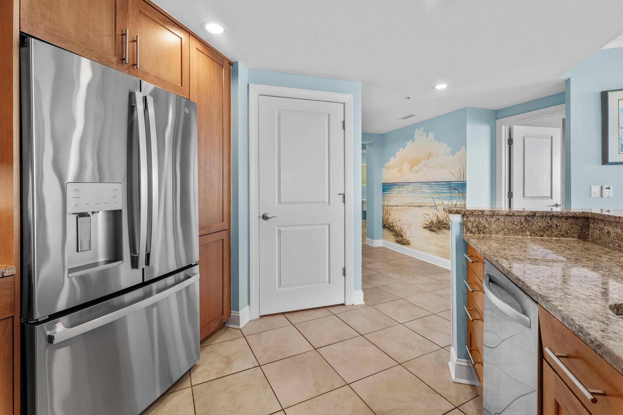 A kitchen with a stainless steel refrigerator, wooden cabinets, a granite countertop, and a door leading to a room with a beach mural.