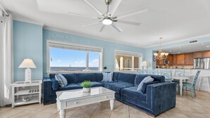 Ein Wohnzimmer mit einem blauen Sofa, einem weißen Couchtisch, einem Deckenventilator und Meerblick durch große Fenster.