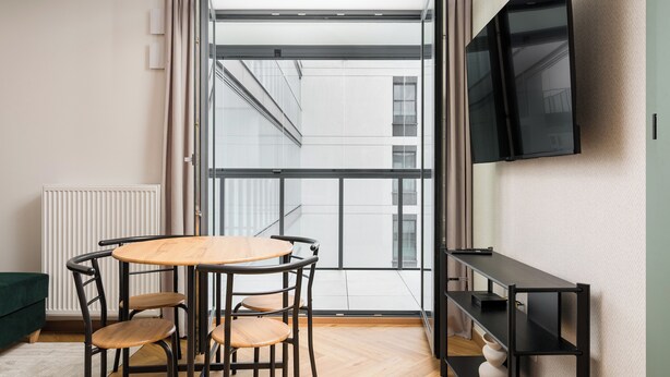 Een moderne kamer met een groot raam, een ronde tafel met stoelen, een zwarte televisie aan de muur en een groene bank.