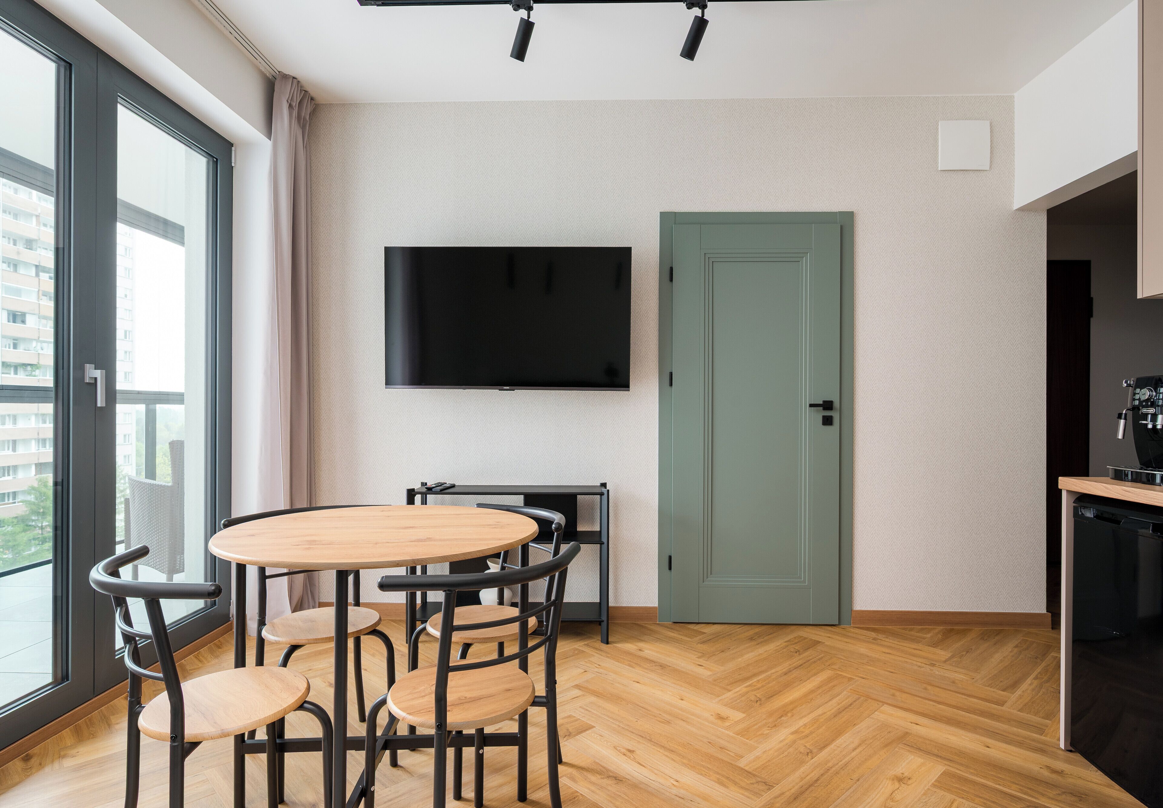 A modern kitchen with a dining area, a television mounted on the wall, and a view through the window.