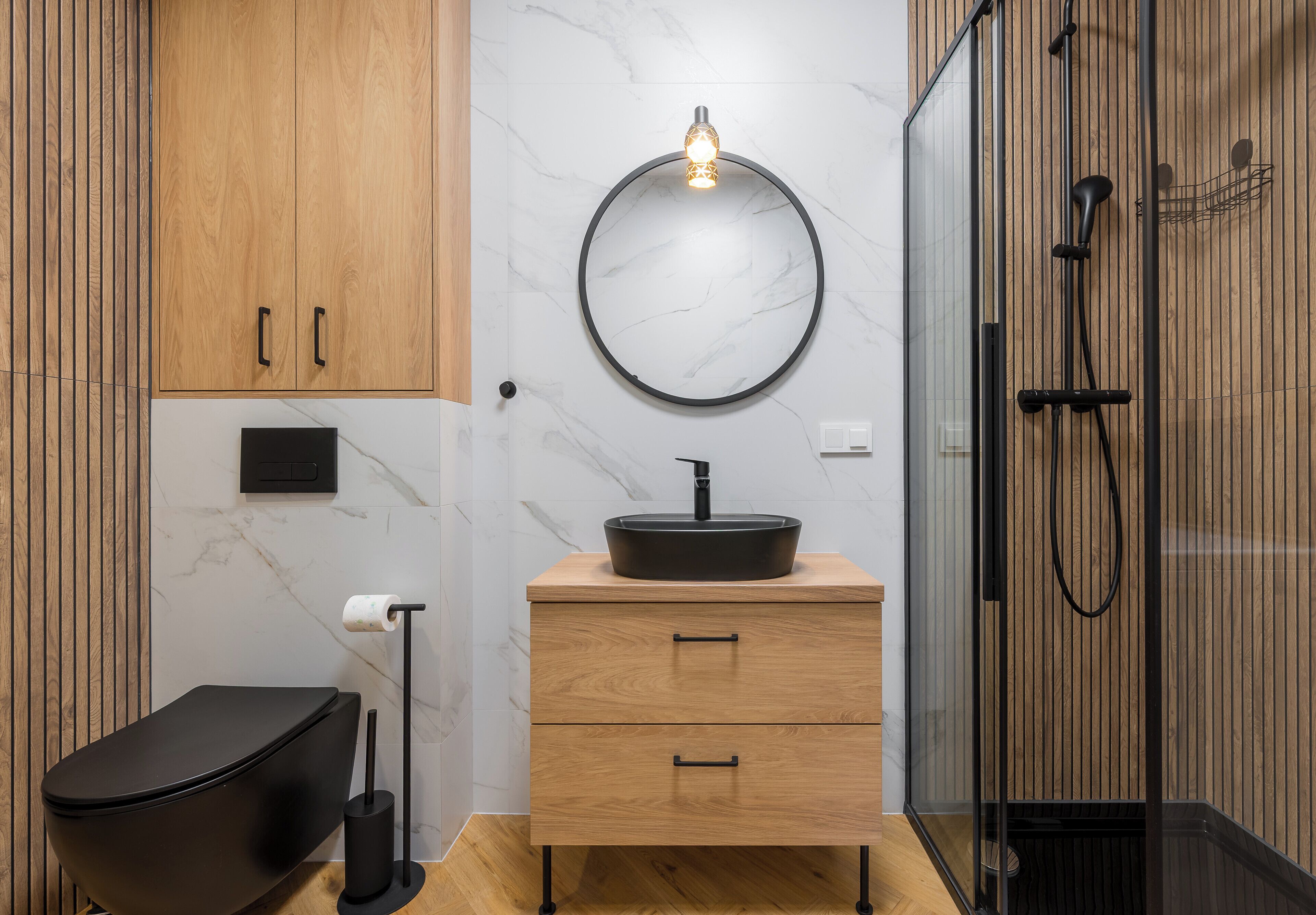A modern bathroom with a black toilet, wooden vanity with a black sink, a round mirror, and a glass shower enclosure.