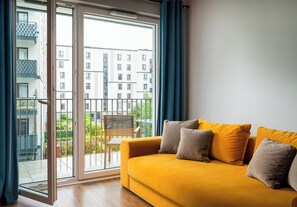En gul soffa med gråa kuddar, en glasdörr som leder ut till en balkong med utsikt över en byggnad och grönska.