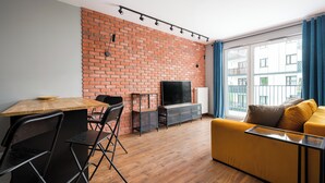 A living room with a brick wall, a TV, a yellow sofa, and a dining area with chairs and a table. - Żerań Brick Loft Apartment (Warszawa)