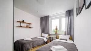 Hotellihuone, jossa on kaksi sänkyä, työpöytä, tuoli, ikkuna, jossa on verhot, ja hylly, jonka päällä on kori.