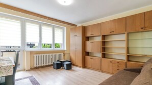A living room with a sofa, a dining table with chairs, a window with blinds, wooden cabinets, and a radiator. - Chorzów Comfort Apartment (Chorzów)