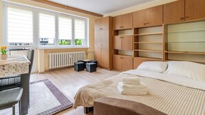 A bedroom with a bed, a desk with a chair, a radiator, and a window with blinds. - Chorzów Comfort Apartment (Chorzów)