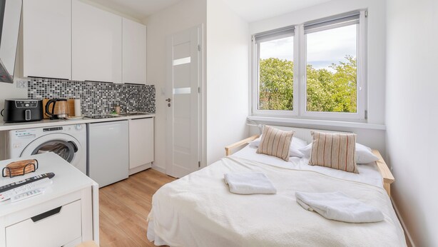 Quarto com cama, almofadas, janela com vista, máquina de lavar roupa e cozinha compacta.