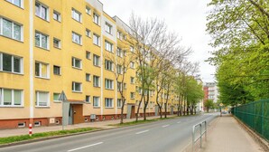 Um edifício amarelo de vários andares com calçada, árvores e uma rua.