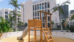 A playground with a slide and climbing structure in an urban setting with modern buildings in the background.