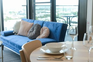 A blue sofa with cushions, a dining table with a bowl and wine glass, and a view through large windows.