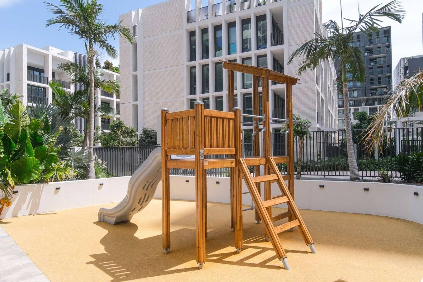 A playground with a slide and climbing structure, surrounded by palm trees and modern buildings.