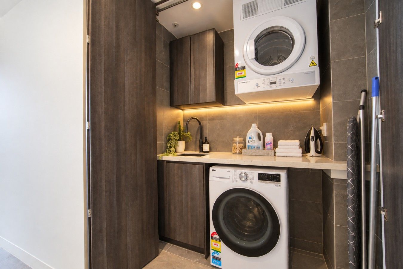 A compact laundry room with a front-loading washing machine, a sink, and a countertop with various items.